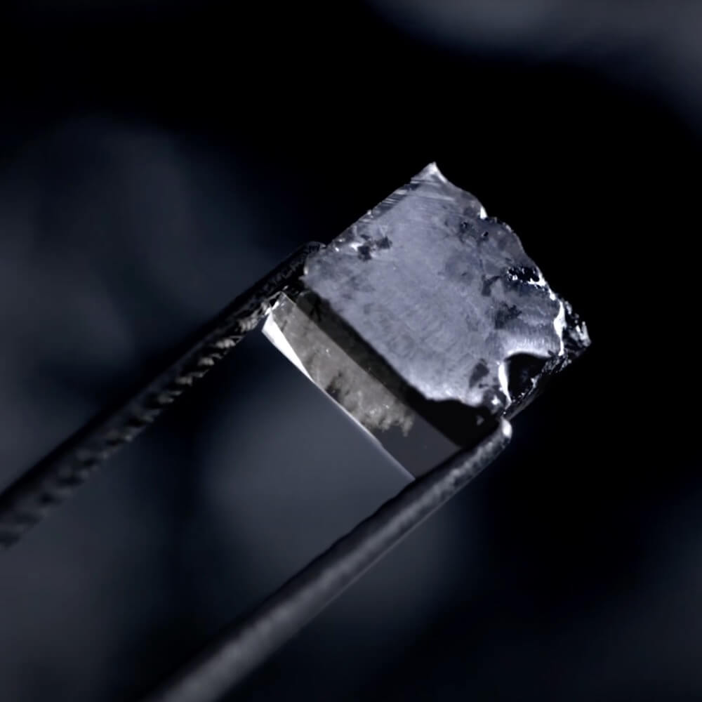 Rå laboratorieskabt diamant holdt i tang mod mørk baggrund.