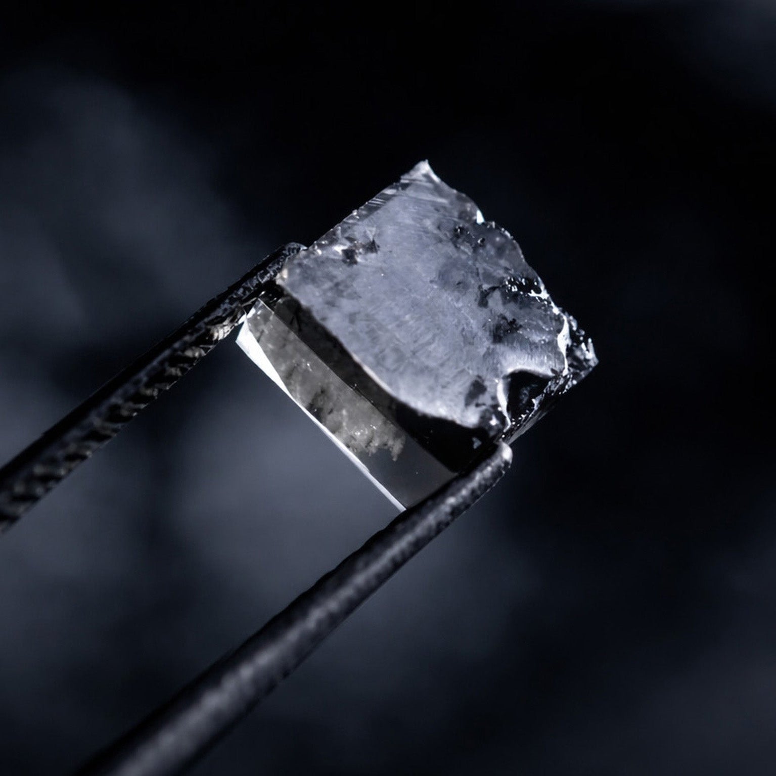 Rå laboratorieskabt diamant holdt i tang mod mørk baggrund.