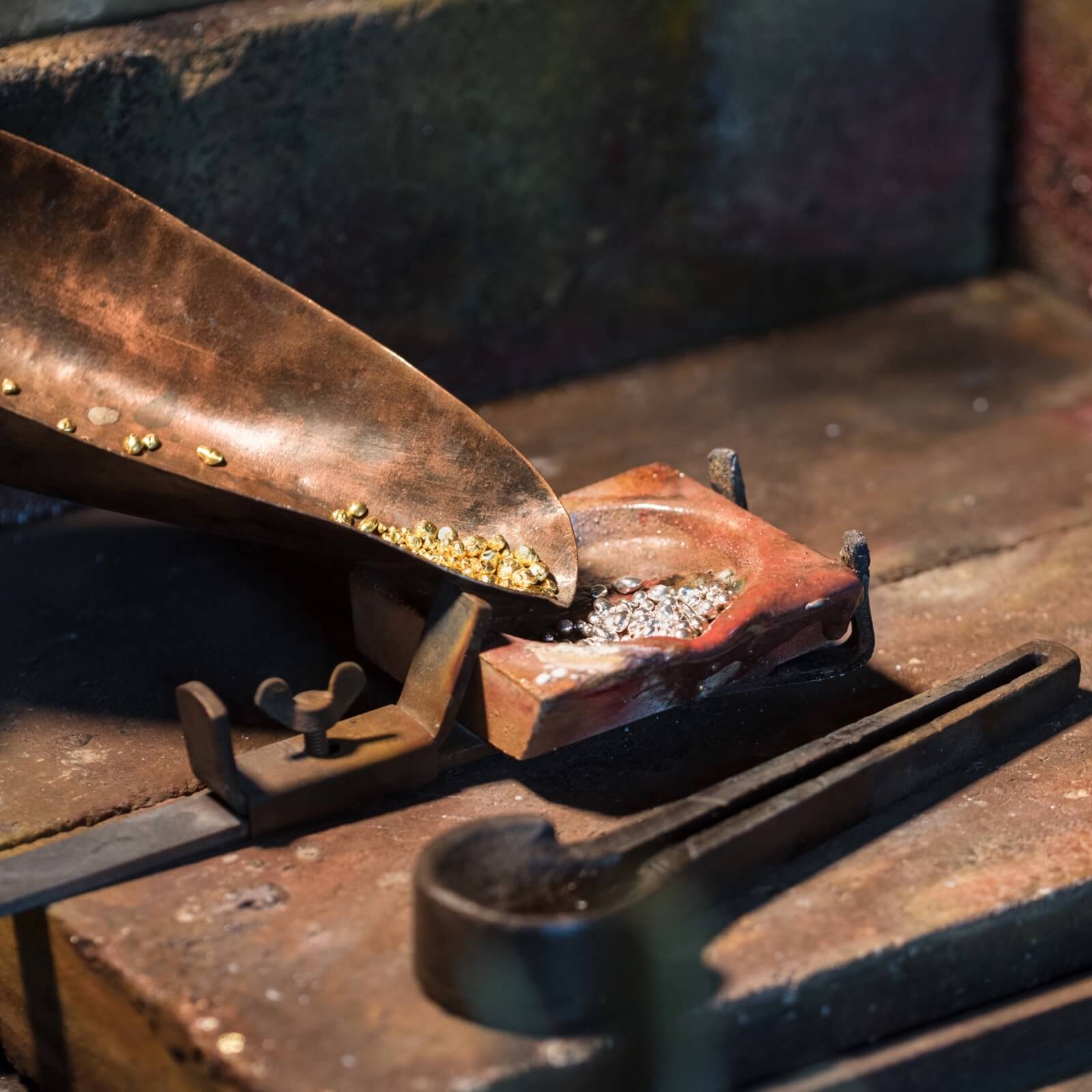 Guld- og sølvgranulater i metalskovl og smelteform på guldsmedens arbejdsbord.