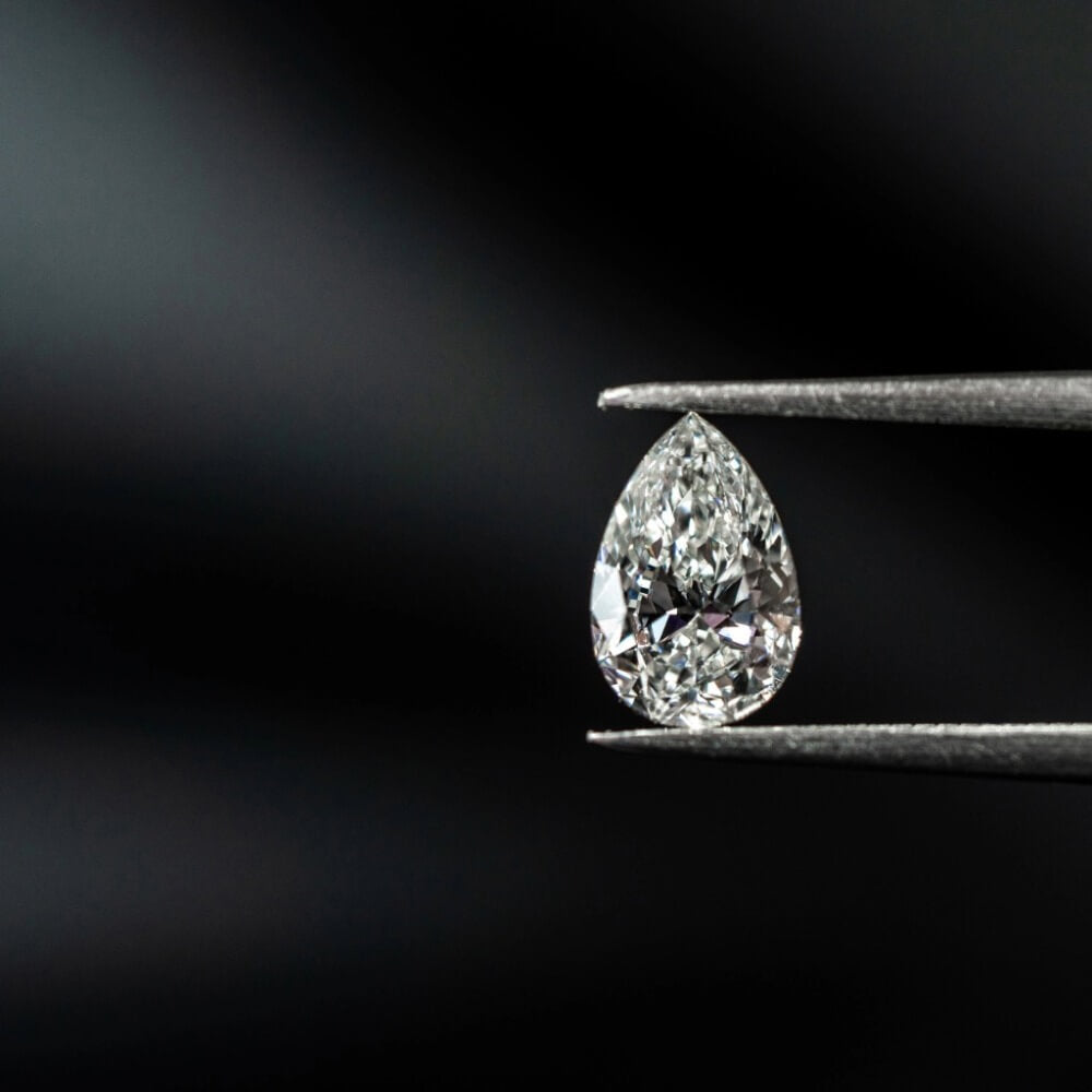 Pæreformet poleret diamant holdt i guldsmedens tang mod mørk baggrund.