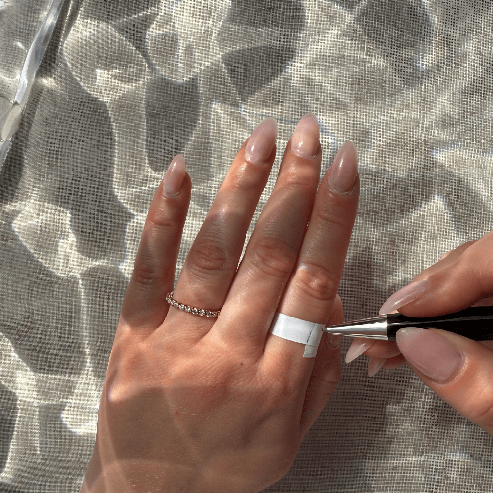 Hånd der måler ringstørrelse med papirsstrimmel og pen i naturligt lys.