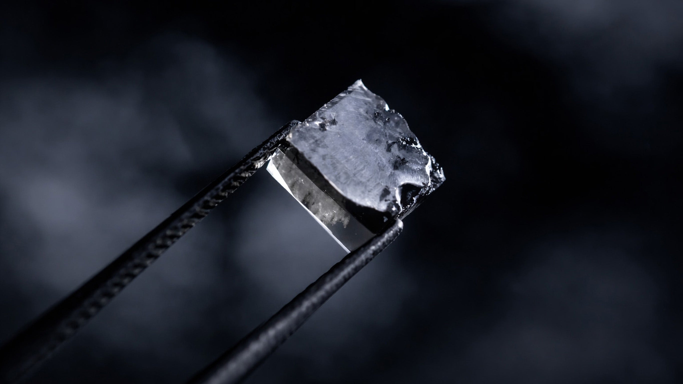 Rå laboratorieskabt diamant holdt i tang mod mørk baggrund.