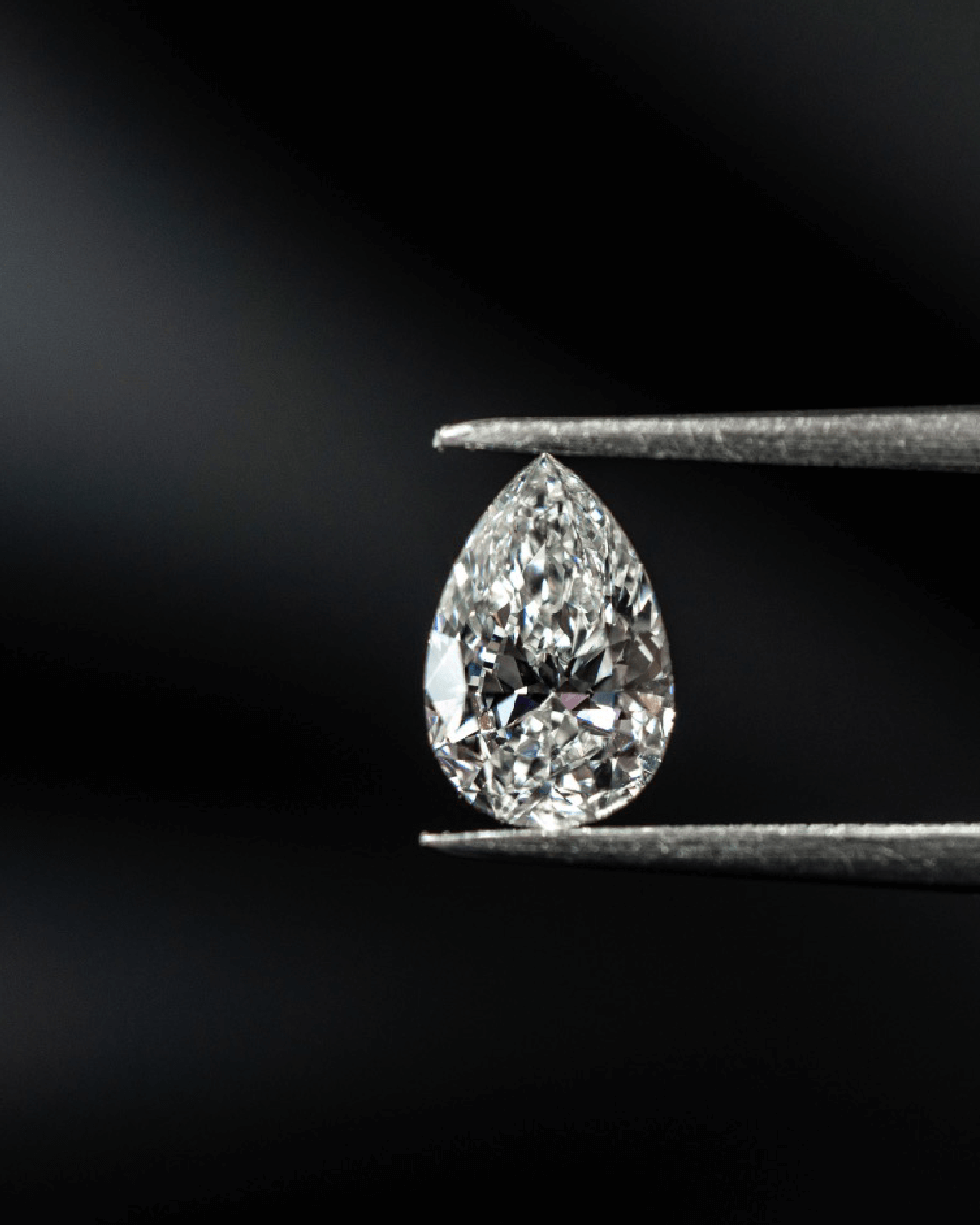 Pæreformet poleret diamant holdt i guldsmedens tang mod mørk baggrund.