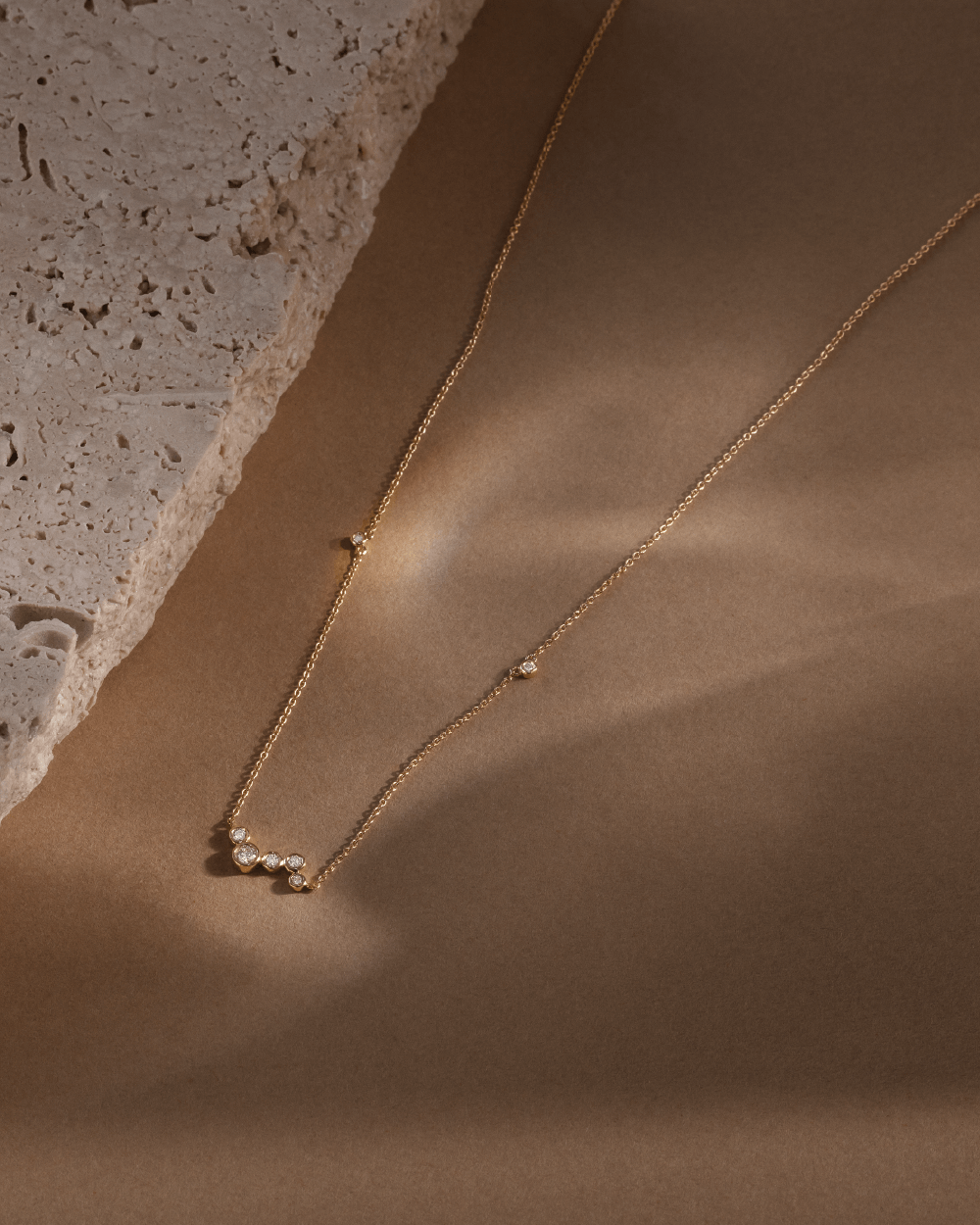 Softpack foto med Eclipse Cluster halskæde i 14 kt guld på brun baggrund med travertin sten ved siden af.