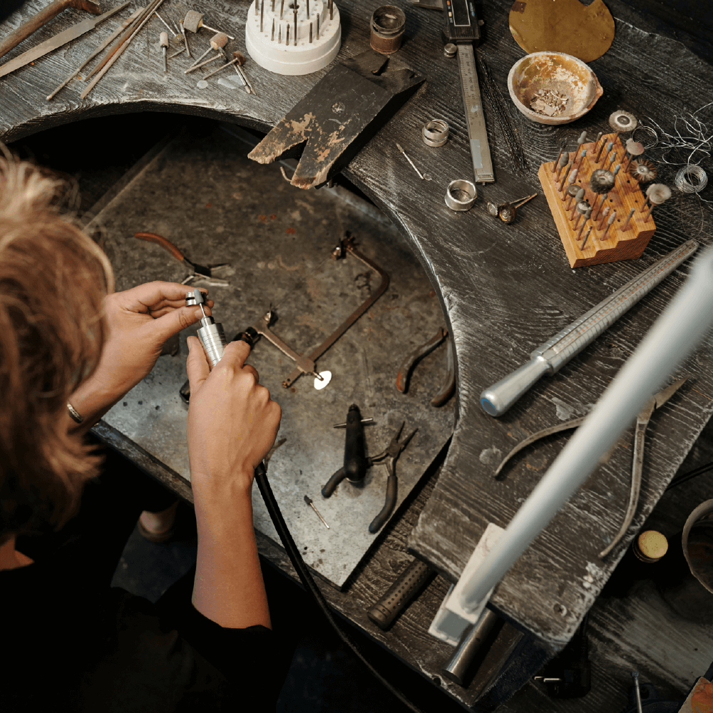 Guldsmed, der arbejder ved et værkstedsbord omgivet af værktøj, måleinstrumenter og smykkekomponenter.