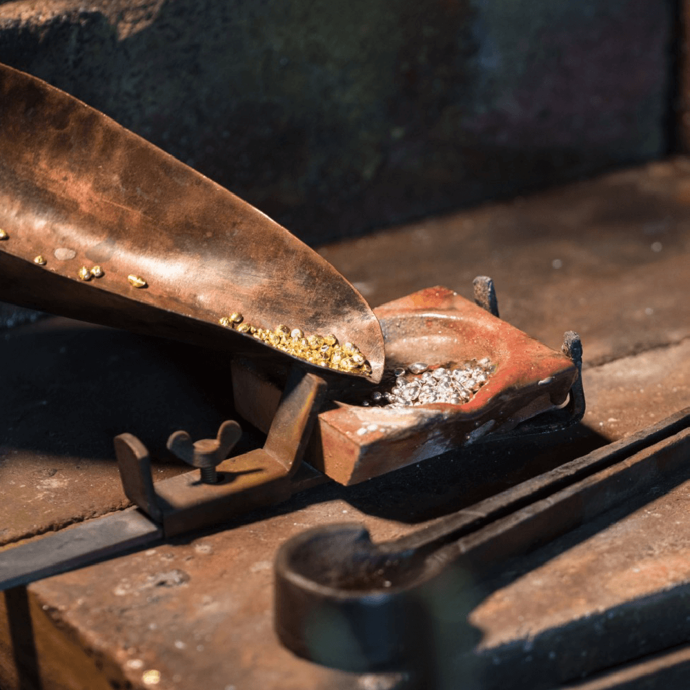 Guld- og sølvgranulater i metalskovl og smelteform på guldsmedens arbejdsbord.