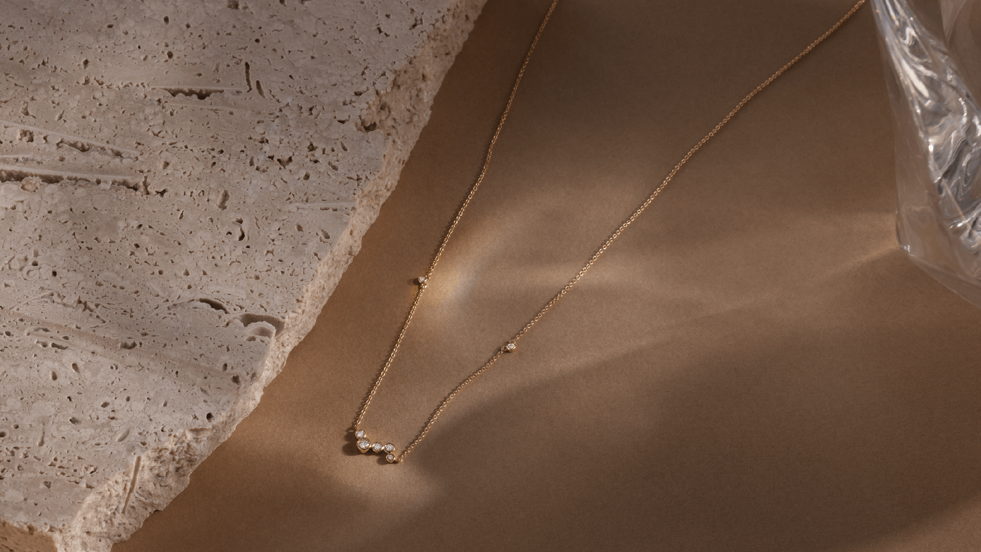 Softpack foto med Eclipse Cluster halskæde i 14 kt guld på brun baggrund med travertin sten ved siden af.